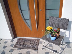 Una porta in legno con un vetrino, un tappetino d'ingresso, una sedia con seduta intrecciata e piante in vaso con fiori gialli.