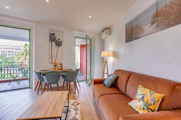 Ein modernes Wohnzimmer mit einer braunen Couch, einem Esstisch mit Stühlen, einem großen Fenster und Blick ins Freie.