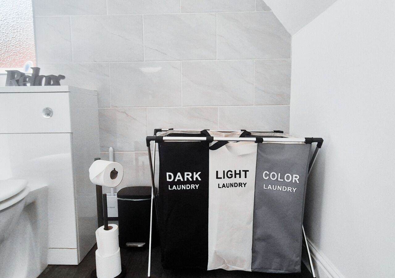 Ein Badezimmer mit Toilette, einem Mülleimer und drei Wäschebeuteln mit den Aufschriften 'DUNKLE WASCHE', 'LICHTE WASCHE' und 'FARBWEASCHE'.