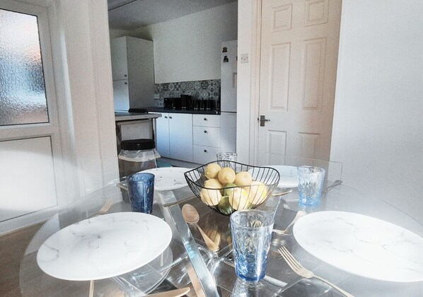 A dining area with a glass table, white plates, a bowl of fruit, and blue glasses. - Cosy 2BR House on Quiet Street (Nottingham)