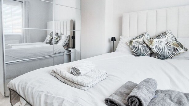 A modern hotel room with a large mirror, a bed with white linens, and a nightstand with a lamp. - Cosy 2BR House on Quiet Street (Nottingham)