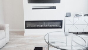 A modern living room with a glass-top coffee table, a flat-screen TV mounted above a fireplace, and a white cabinet with a geometric tile design. - Cosy 2BR House on Quiet Street (Nottingham)