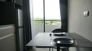 A dining area with a table set for two, a view of greenery and mountains through a large window, and a modern kitchen with a refrigerator and cabinets. - Palm Bay Studio T3 -532 (Chalong)