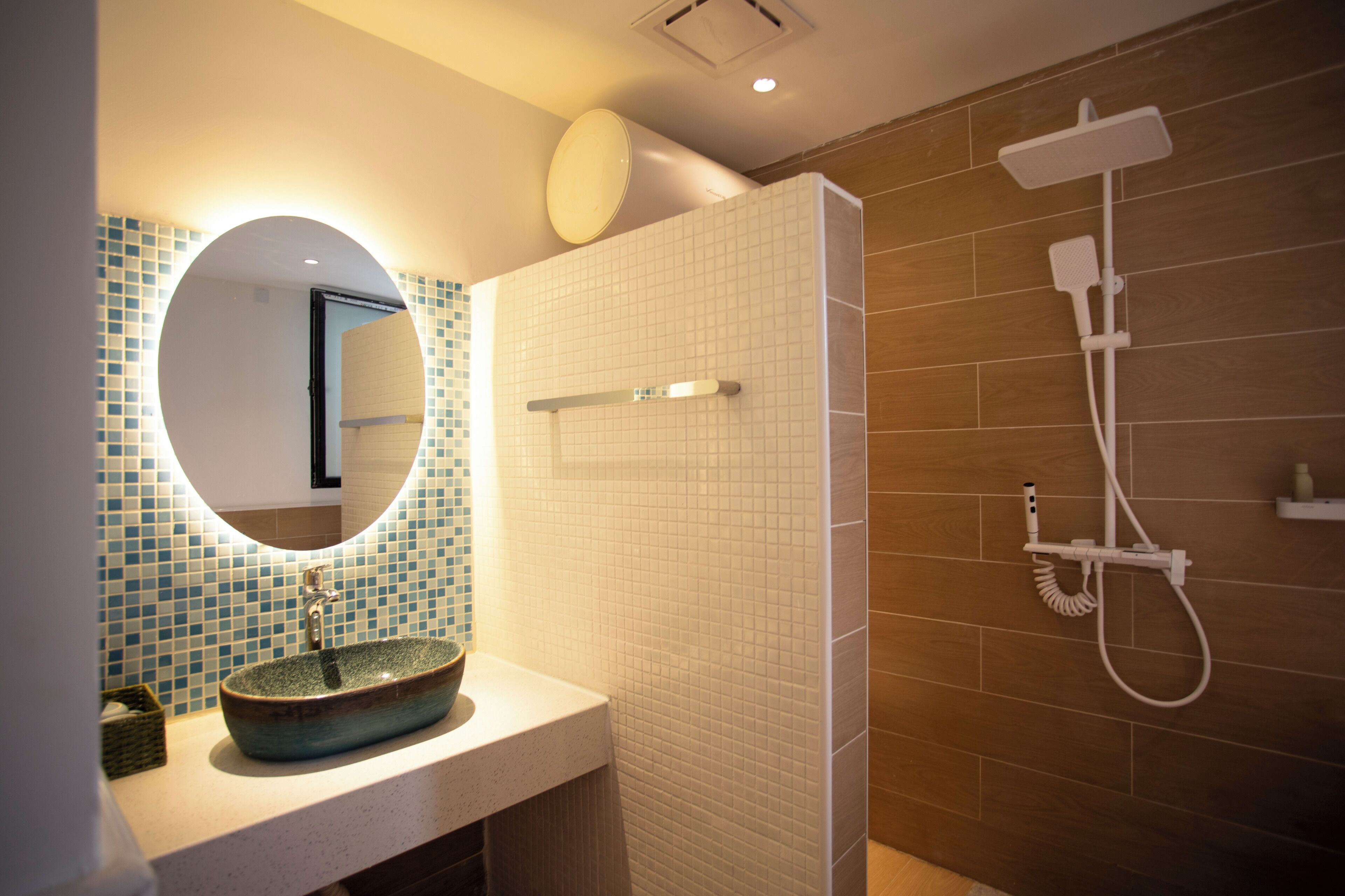 A bathroom with a round mirror, a vessel sink, a shower with a handheld showerhead, and a tiled wall.