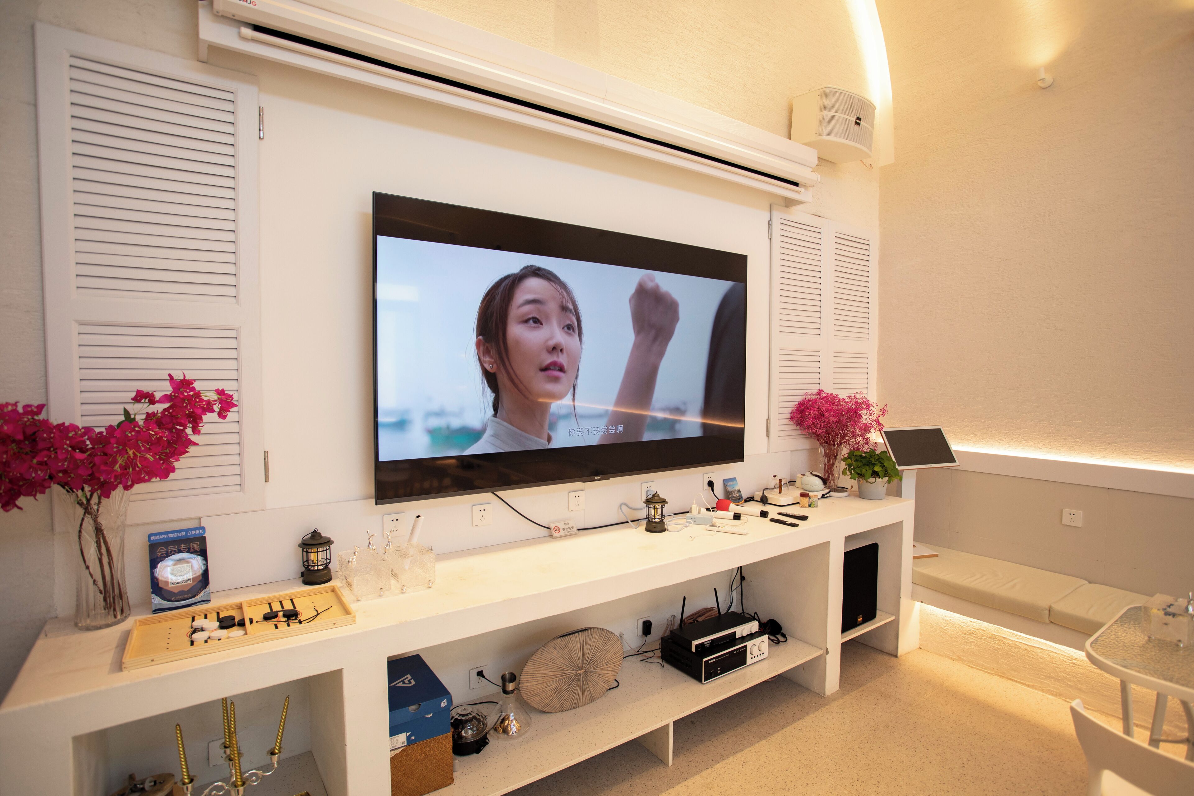 A modern living room with a flat-screen TV mounted on the wall, a white entertainment unit below, and a glass table with chairs.