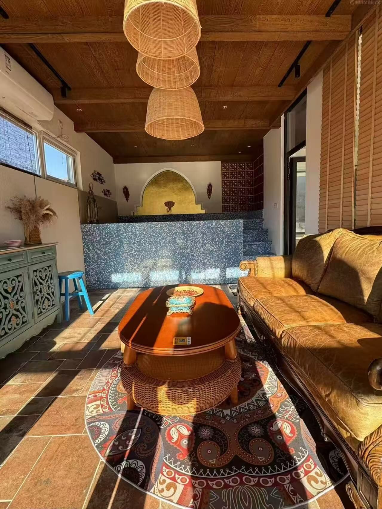 Quarto com uma mesa de centro redonda de madeira, um sofá, um tapete estampado e um painel de azulejos azuis.