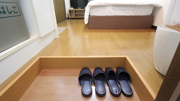 A hotel room with a wooden floor, a bed with white bedding, a television on a stand, and a shoe rack with black slippers. - Small-Town Feel, Big-City Access | Homey Stay in Nakano (Nogata, Nakano-ku)