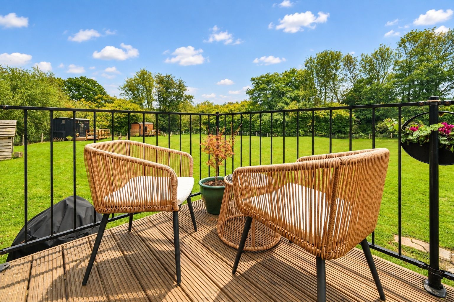 Une terrasse avec deux fauteuils en osier et une table, entourée d’une clôture en métal noir, donnant sur une pelouse verdoyante et des arbres.