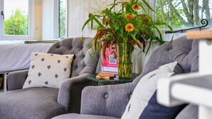 Ett mysigt vardagsrum med en grå soffa, ett litet bord med en vas med blommor och ett fönster med utsikt över träd.