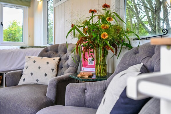 A cozy living room with a grey sofa, a small table with a vase of flowers, and a window with a view of trees. - Badger Shepherds Hut - a holiday let that sleeps 2 guests in 1 bedroom (Hartfield)