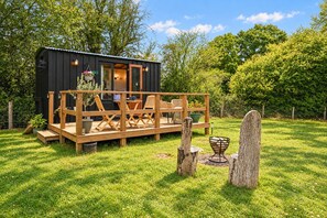 Una terraza de madera con mobiliario exterior, un edificio pequeño con una puerta y un jardín con árboles y una cerca.
