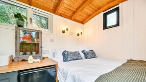 A bedroom with a wooden ceiling, a bed with white bedding and patterned pillows, a small window, and a wooden desk with a refrigerator.