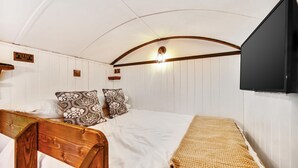 A bedroom with a wooden headboard, a bed with white bedding, patterned pillows, a beige throw blanket, a wall-mounted TV, and a light fixture. - Stag Shepherds Hut -  a holiday let that sleeps 4 guests  in 1 bedroom (Hartfield)