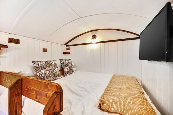 Une chambre à coucher avec une tête de lit en bois, un lit avec une literie blanche, des coussins à motifs, un plaid beige, un téléviseur fixé au mur et un luminaire.