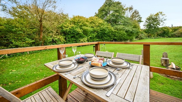 A wooden deck with a table set for a meal, surrounded by chairs, overlooking a grassy area with trees. - Stag Shepherds Hut -  a holiday let that sleeps 4 guests  in 1 bedroom (Hartfield)