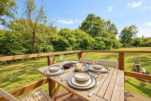 An outdoor dining area with a wooden deck, set tables with plates, cutlery, and a bowl of fruit.