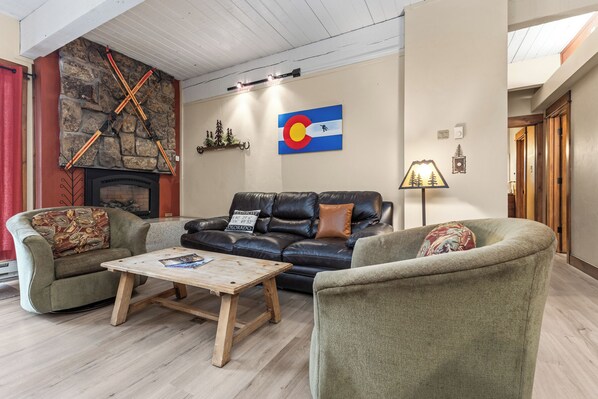 Una sala de estar acogedora con chimenea, un sofá de cuero negro, dos sillones verdes y una mesa de centro de madera.
