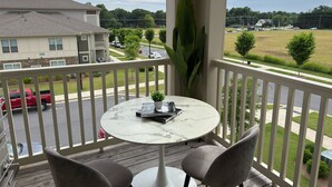 Een balkon met een ronde tafel en twee stoelen, met uitzicht op een straat met geparkeerde auto's en een woongebouw.