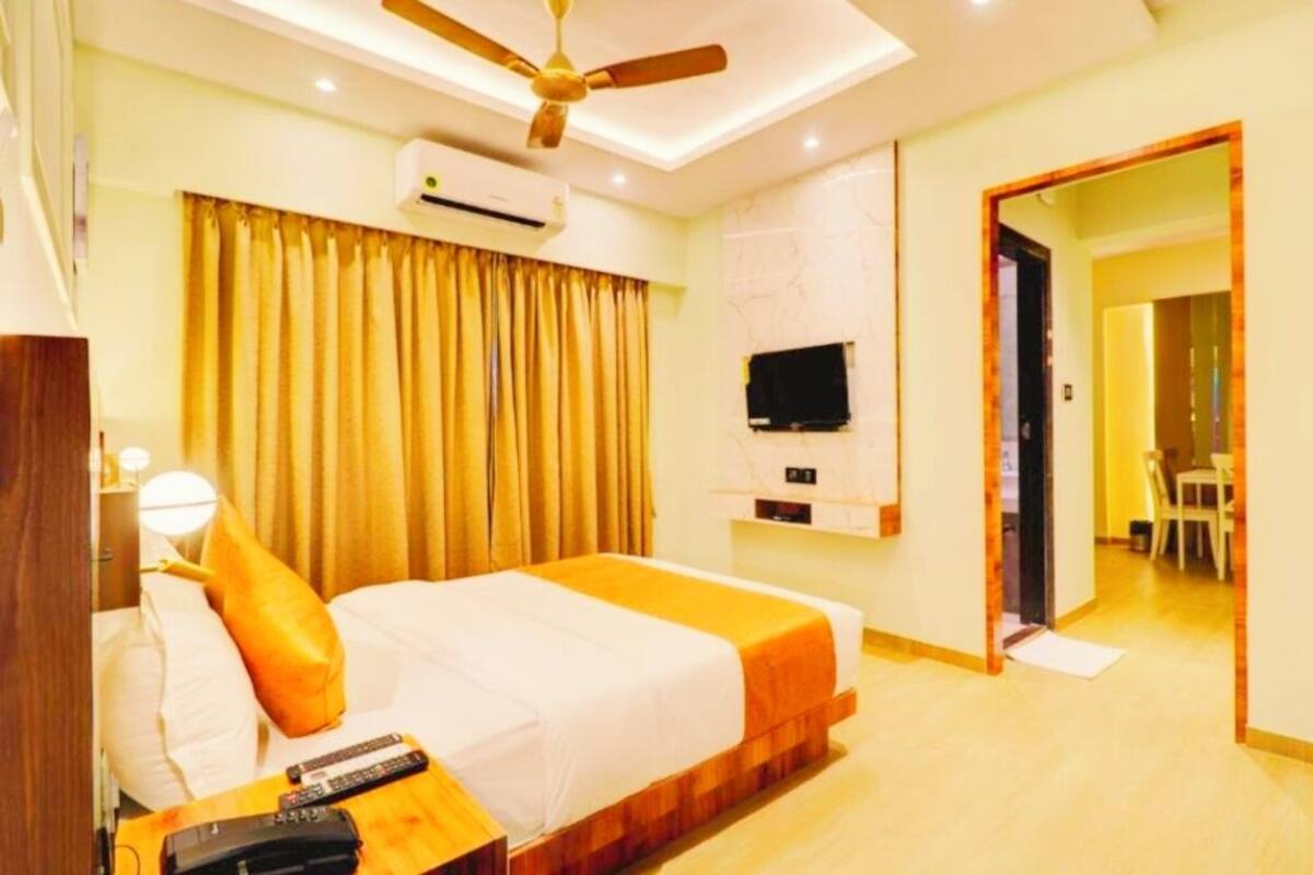 A hotel room with a bed, a ceiling fan, an air conditioning unit, a television, and a dining area visible through an open door.