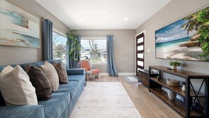 Een woonkamer met een blauwe bank, een televisie op een houten dressoir en een groot schilderij van een strand.