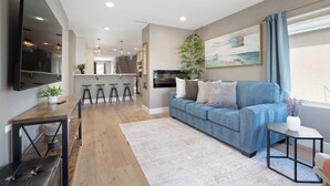 Een moderne woonkamer met een blauwe bank, een flatscreen televisie, een bijzettafeltje en een eethoek op de achtergrond.