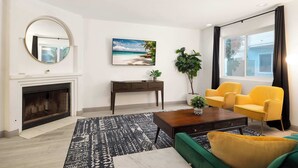 Un salon avec une cheminée, un tableau représentant une plage, un miroir rond et des chaises jaunes.