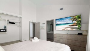 Une chambre avec un grand lit, un téléviseur fixé au mur, une armoire et une salle de bain visible à travers une porte ouverte.