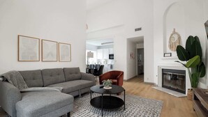 A modern living room with a grey sectional sofa, a round coffee table, and a patterned rug. There is a fireplace with a macrame wall hanging and a large green plant.