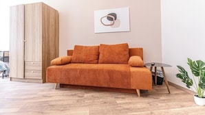 A modern living room with a wooden wardrobe, a sofa with orange cushions, a small round table, and a potted plant. - Chic 1BR Apt Walk to Vienna Sights (Vienna)