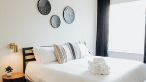 A neatly made bed with white linens, a bedside lamp, and a small potted plant on a round wooden nightstand.