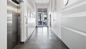 A modern, well-lit hallway with a central door, flanked by elevators and a wall-mounted light fixture.