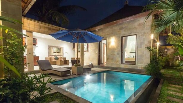 A swimming pool with lounge chairs and a blue umbrella, surrounded by a garden and a building with large windows.