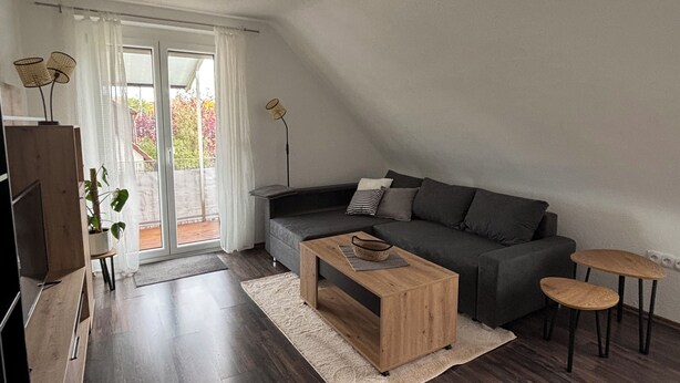 A modern living room with a dark grey sofa, a wooden coffee table, and two small wooden tables. There is a large window with white curtains, a plant, and a floor lamp. - Charming vacation apartment - quiet surroundings but close to the city (Deutschland)