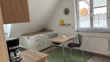 A compact kitchen with a coffee maker, a dining table with chairs, a clock, and a window with curtains.