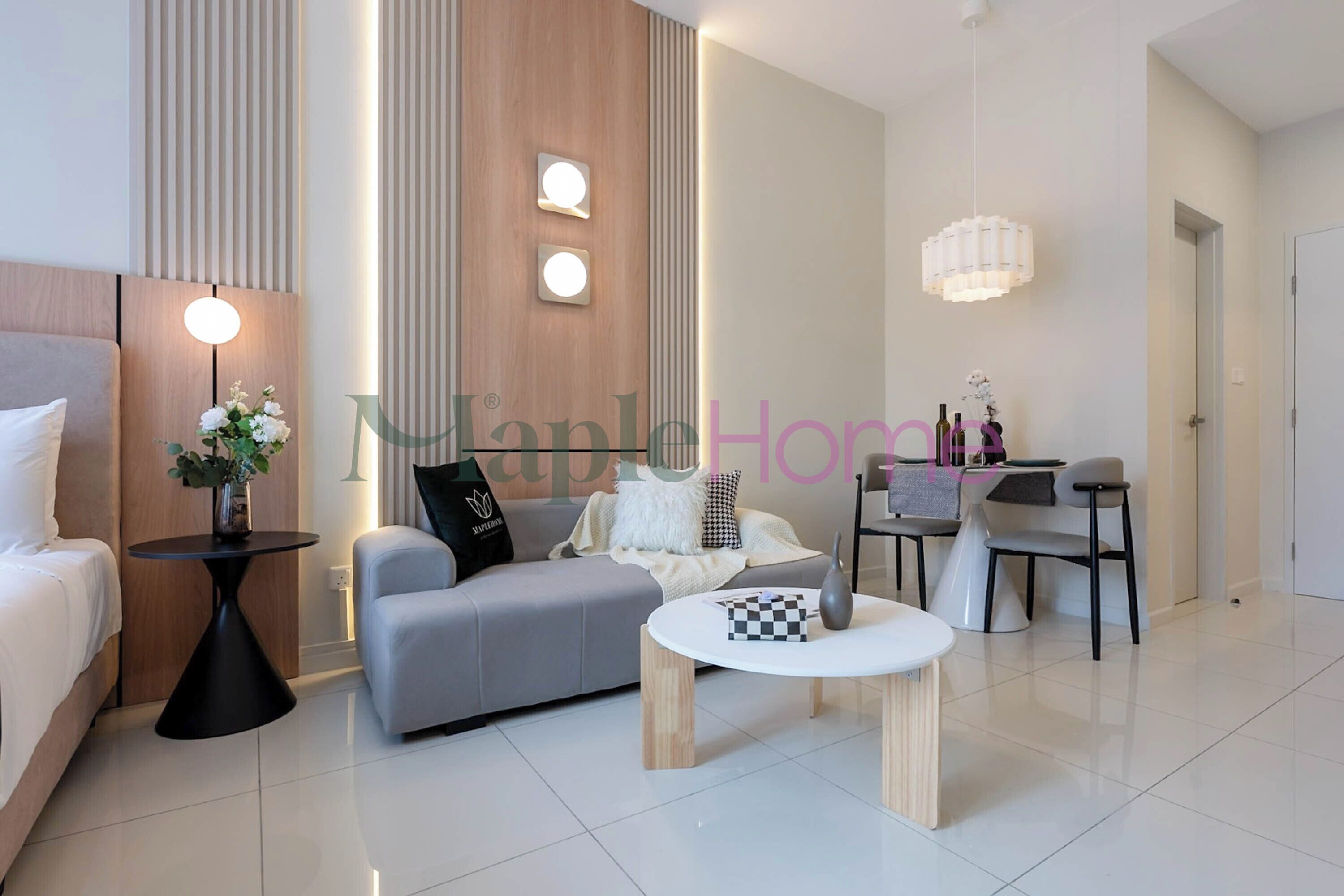 Une chambre d’hôtel moderne comprenant un lit, un canapé gris, une table basse ronde, une petite table d’appoint avec une lampe, et un coin repas avec une table et des chaises.