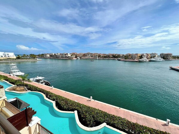 A waterfront view with a swimming pool, a marina with boats, and a row of buildings along the shore.