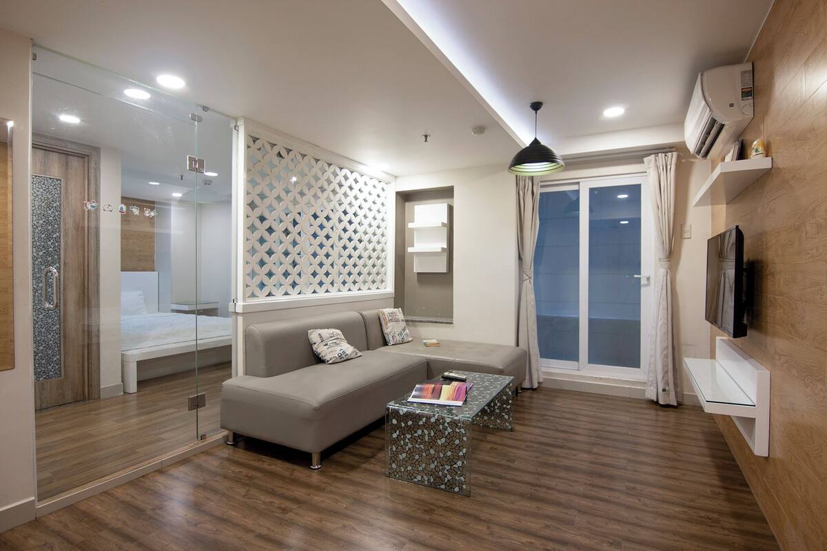 A modern living room with a glass-enclosed shower, a grey sofa with cushions, a patterned coffee table, and a TV on a wooden wall.