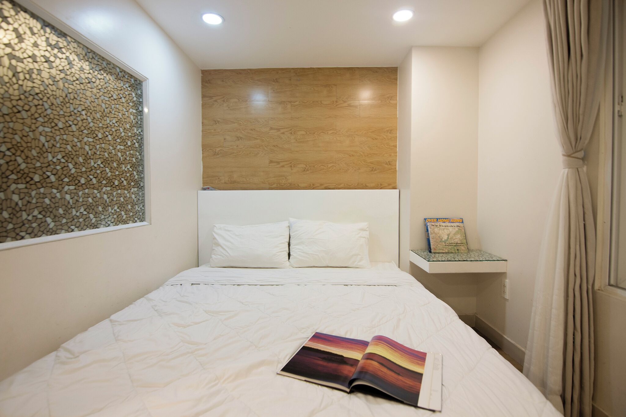 A bedroom with a bed, white bedding, a book on the bed, a wooden headboard, a wall with a textured design, and a small shelf with a book.