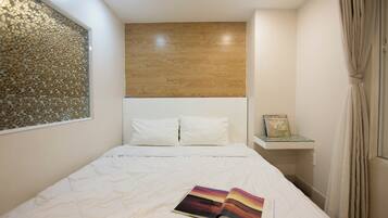 A bedroom with a bed, white bedding, a book on the bed, a wooden headboard, a wall with a textured design, and a small shelf with a book.
