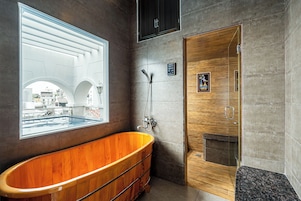 A modern bathroom with a wooden bathtub, a glass-enclosed shower, and a large window offering a view of the cityscape.