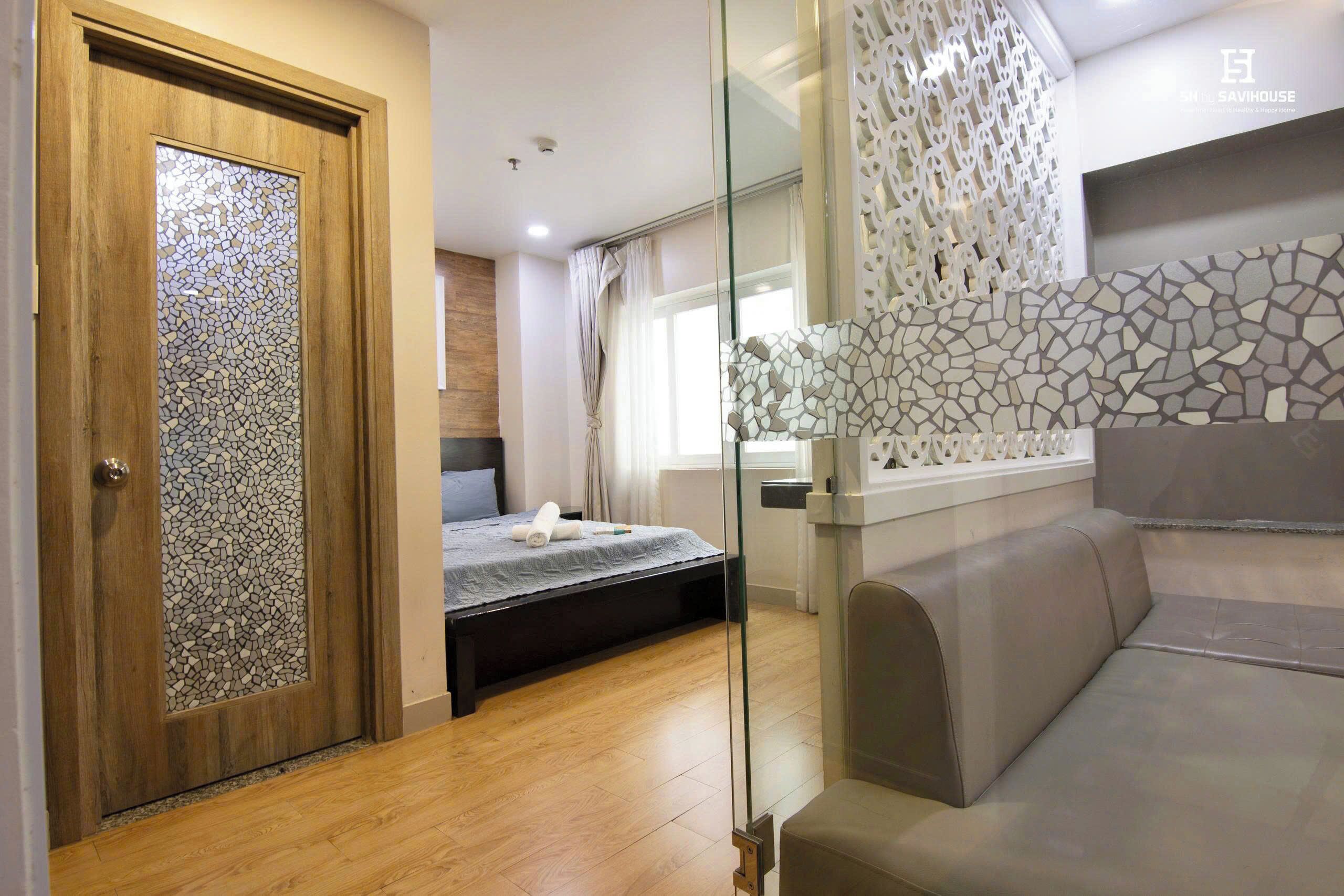 A modern bedroom with a wooden door, a bed with a blue bedspread, a glass partition, and a seating area with a cushioned bench.