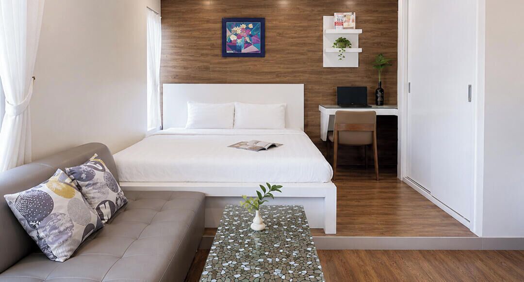 A modern hotel room with a bed, a sofa, a small table with a plant, a desk with a chair, and a wooden wall.
