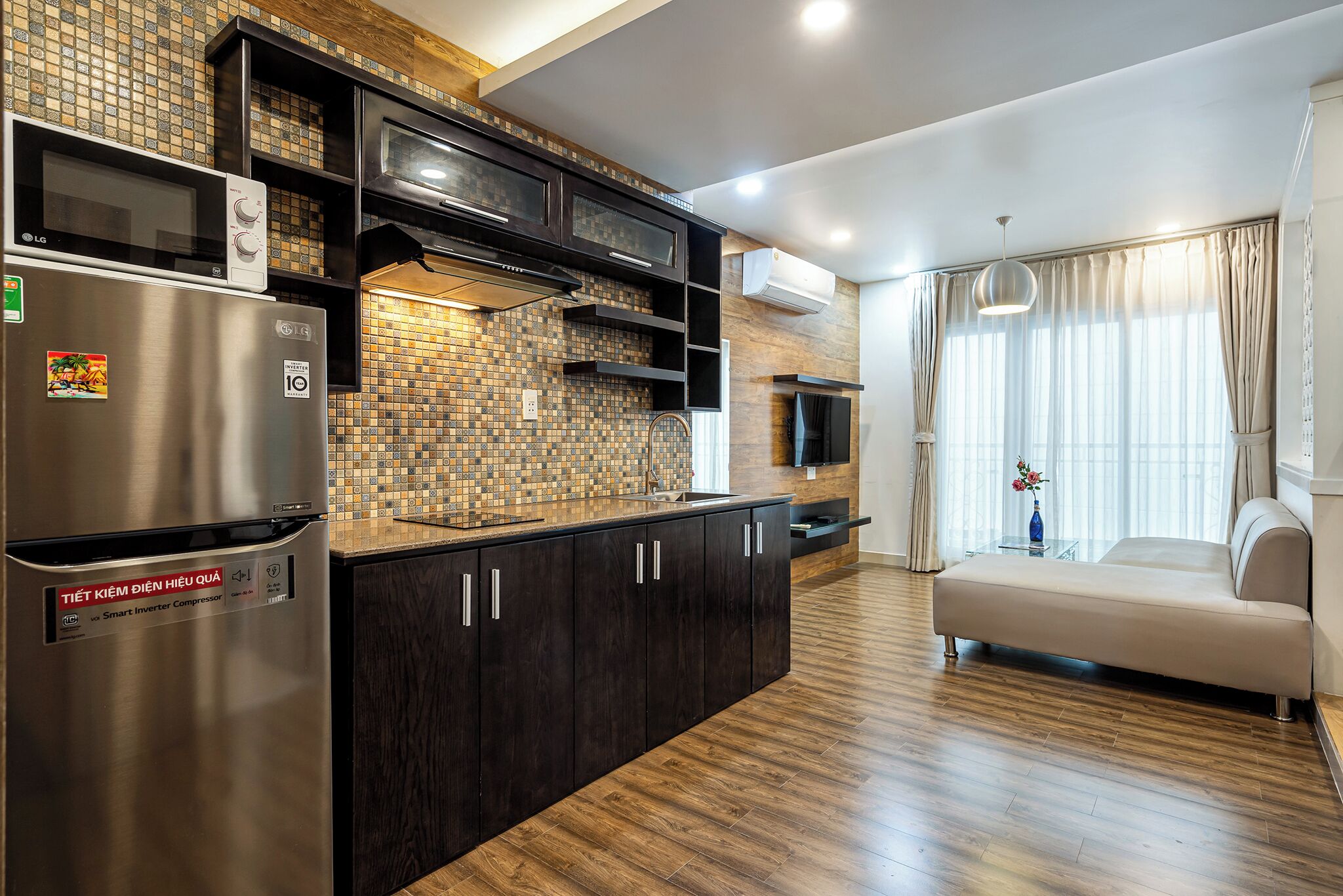 A modern kitchen with dark cabinetry, a stainless steel refrigerator, and a microwave. There is a wooden floor and a beige sofa in the adjacent living area.