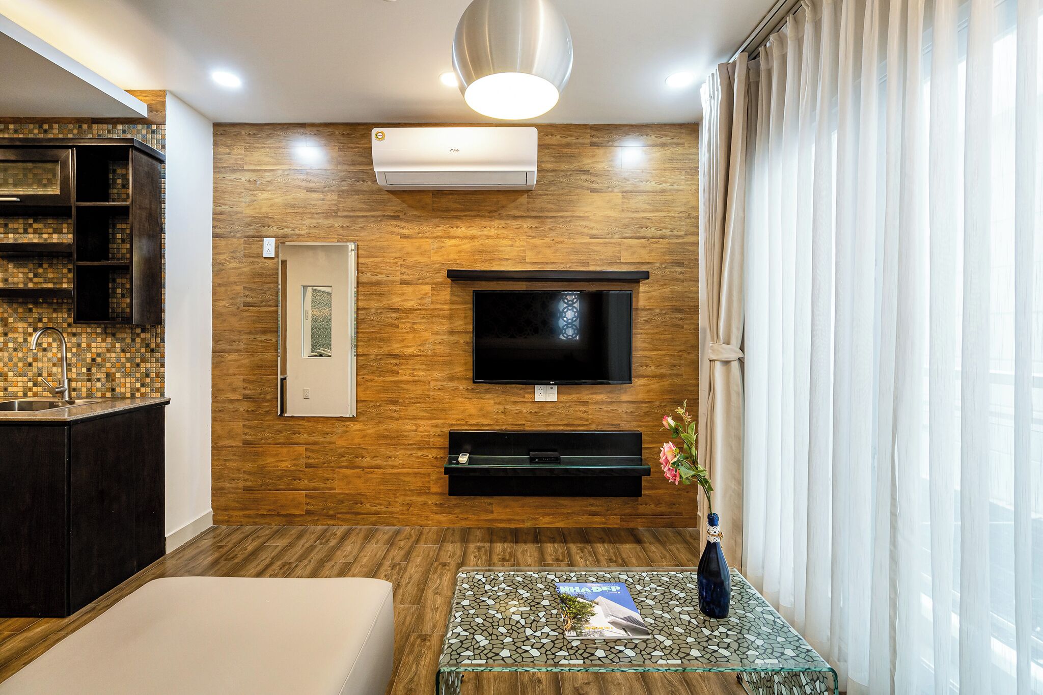 A modern living room with a wooden wall, a mounted TV, a glass coffee table with magazines, and a vase with flowers.
