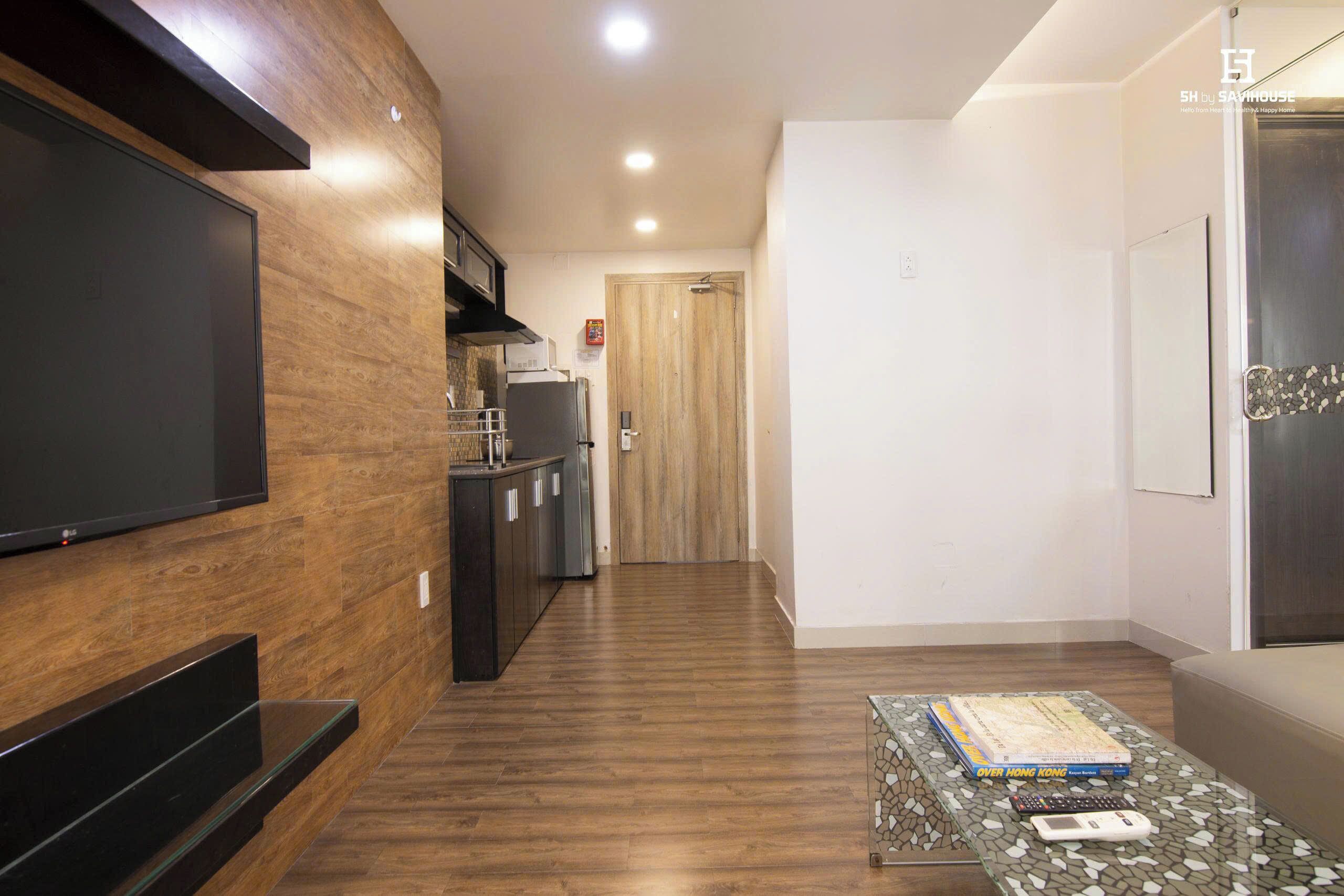 A modern living room with a flat-screen TV, a kitchen area with a refrigerator, and a dining table with books and a remote control.