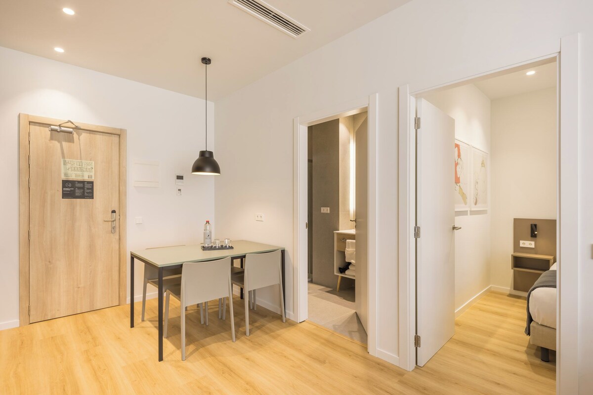 Una habitación moderna y luminosa con suelo de madera, una mesa de comedor con sillas, una lámpara colgante y una puerta que da a un baño.