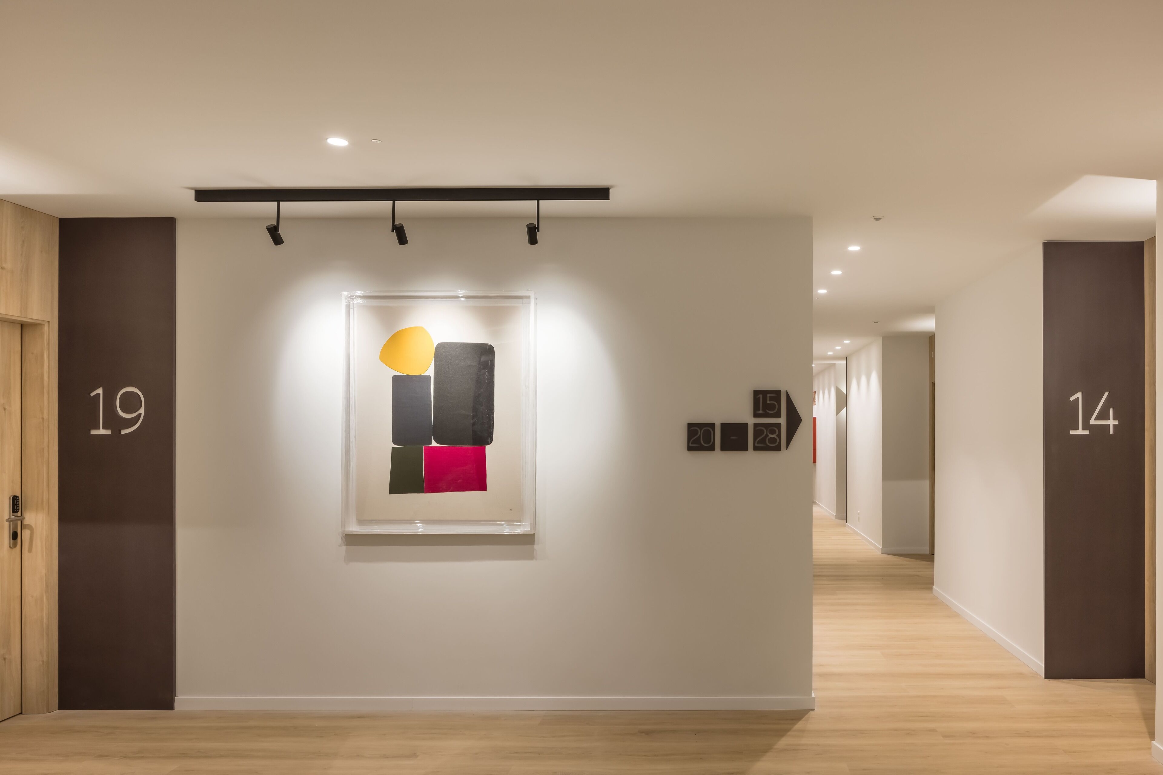 Un couloir avec des portes numérotées, une œuvre d’art abstraite encadrée et un plancher en bois.