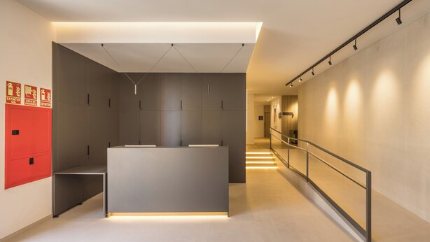 A modern reception area with a minimalist design, featuring a reception desk, a red emergency exit sign, and a staircase leading to another floor. - Mon Suites Jardín de Ayora (València)