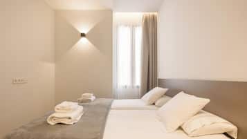 Una habitación de hotel con dos camas, una lámpara fijada a la pared, una ventana con cortinas y una toalla doblada sobre la cama.