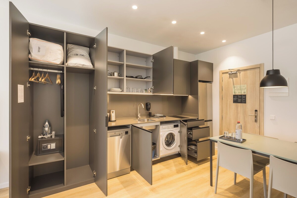 Een kleine keuken met een wasmachine, een koffieapparaat en een eethoek met een tafel en stoelen.